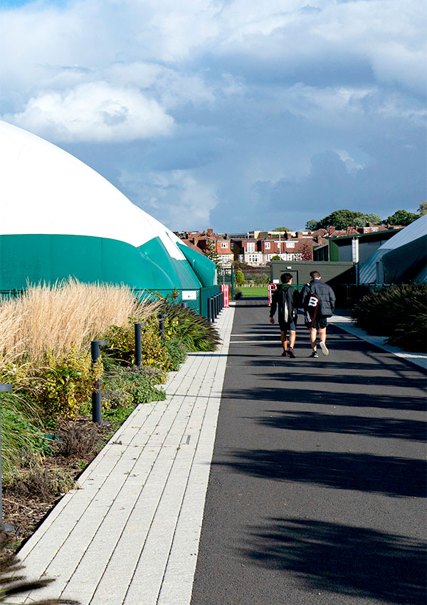 Two tennis players walk along a path lined with plants situated between two airdromes